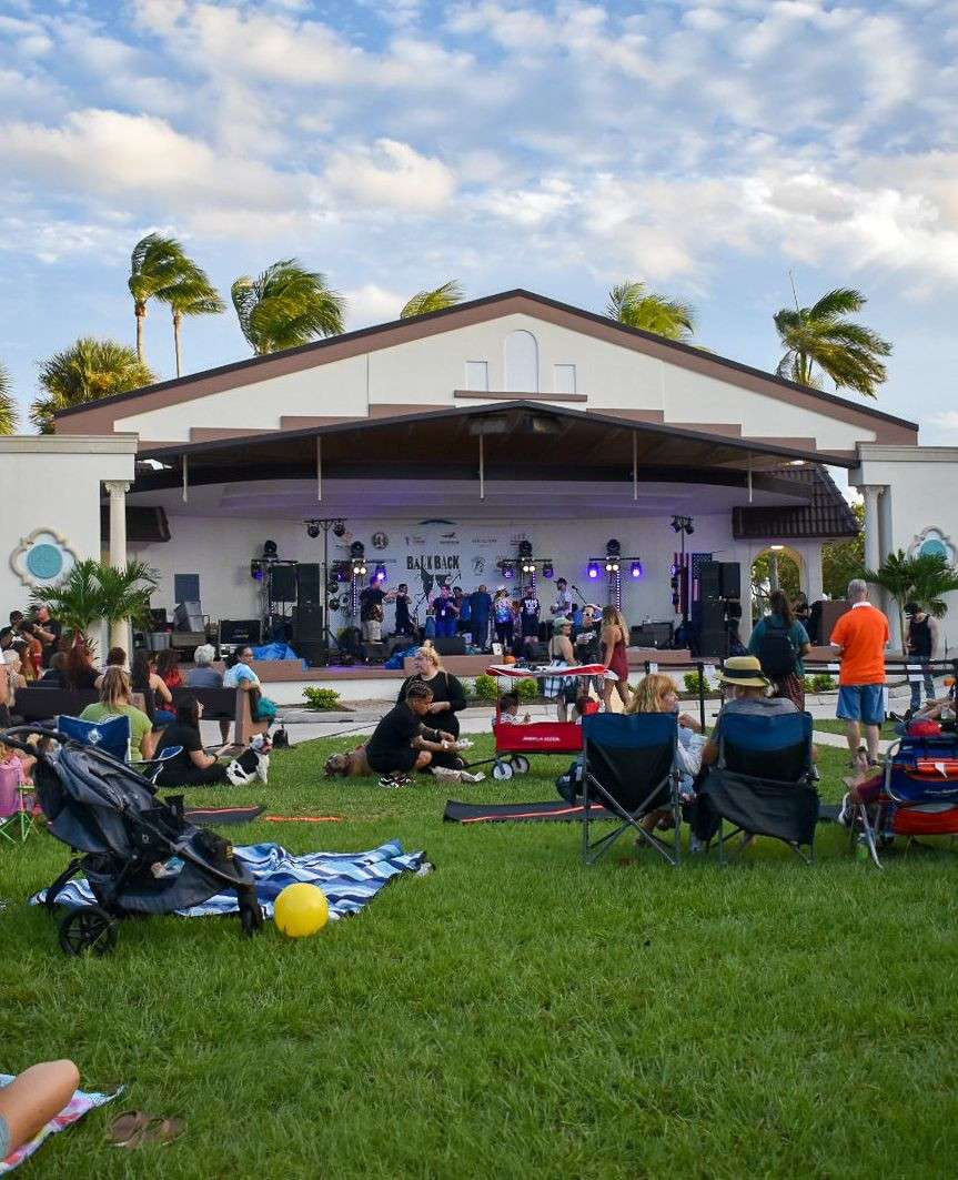 Concierto de música al aire libre con escenario, público y palmeras de fondo.