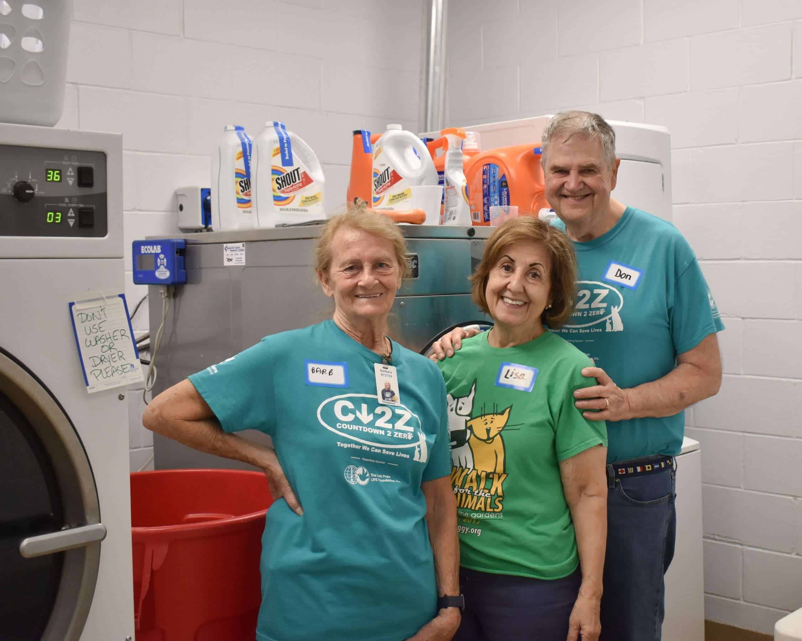Volunteers in teal and green shirts smiling in laundry room with cleaning supplies.