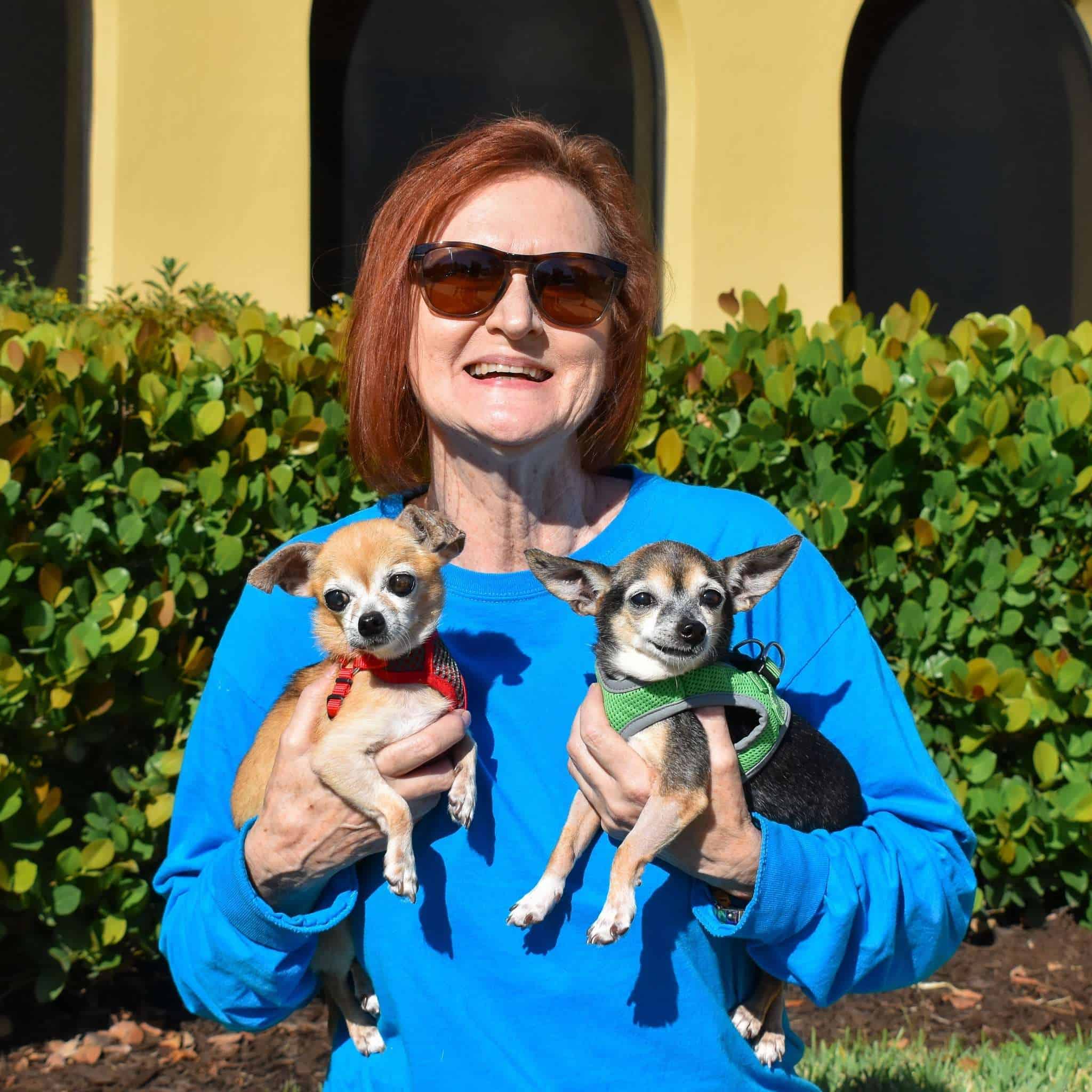 Mujer sosteniendo dos perros pequeños, sonriendo, frente a un edificio amarillo y arbustos verdes.