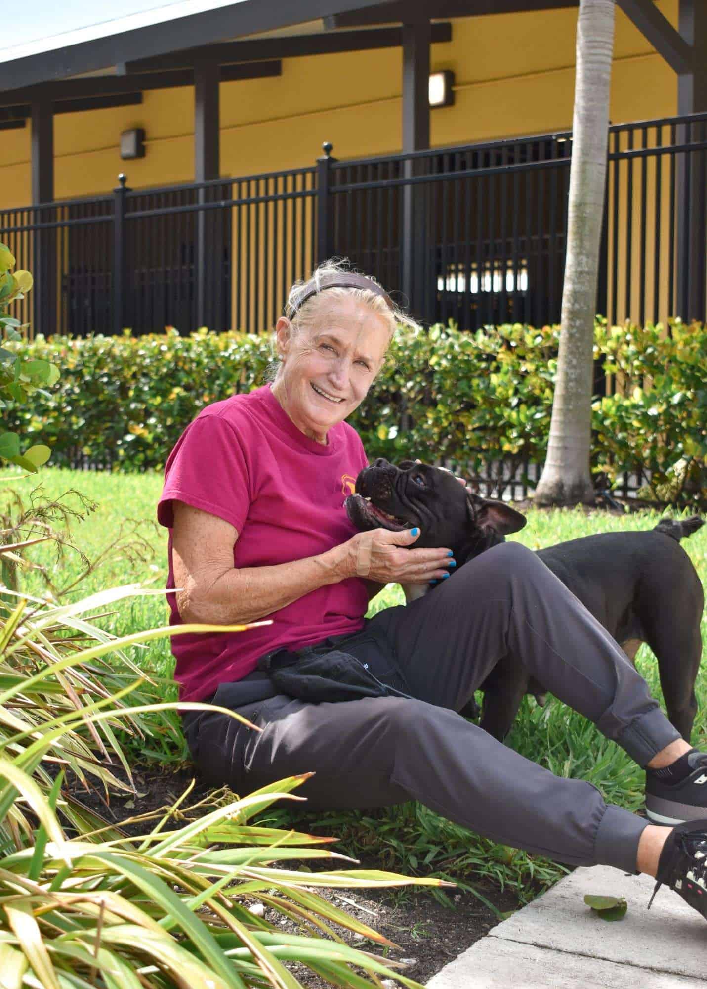 Senior woman smiling with black dog, sitting on grass in pet shelter garden.