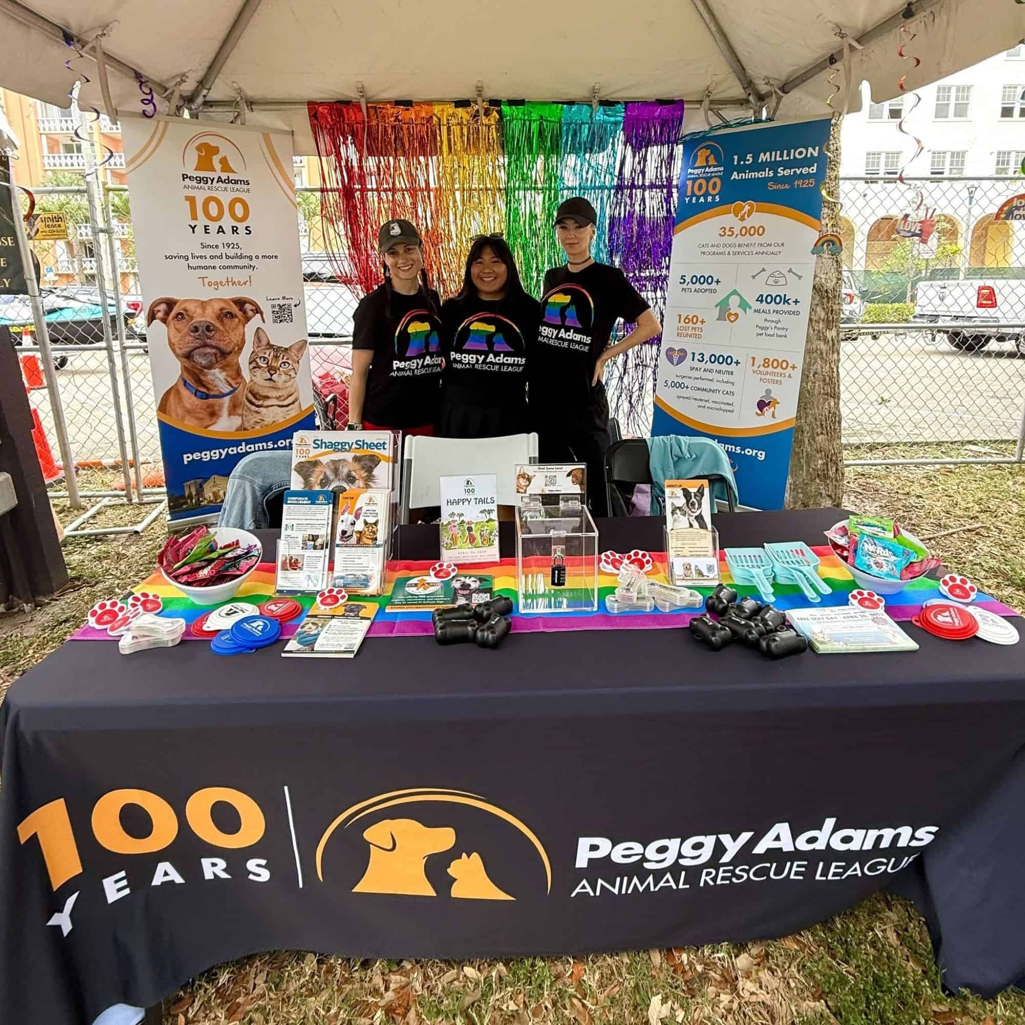 Peggy Adams Animal Rescue League staff and volunteers at a booth with a colorful rainbow backdrop.